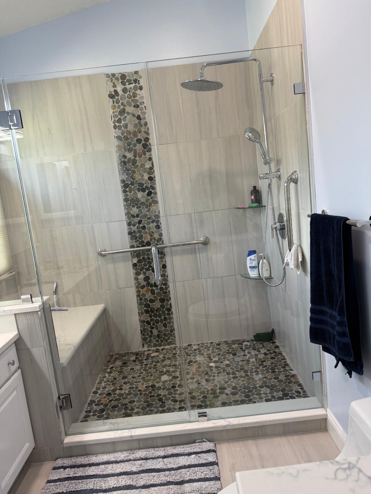 A modern shower with glass doors, stone tile floor and accent, and grab bars.