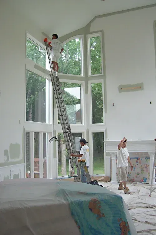 People painting the interior of a house with tall windows. One on a ladder, two at the base.