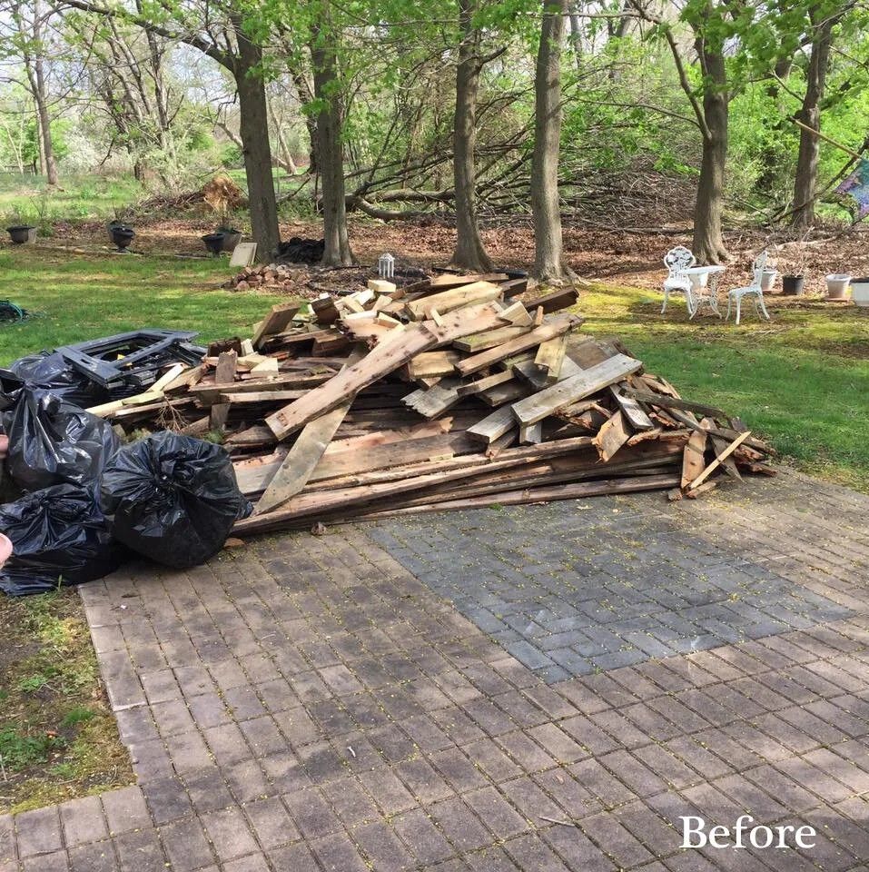 Pile of wooden planks and black trash bags on a brick patio in a backyard with trees.