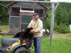 A man is holding a black bear on the back of a lawn mower.