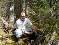 A man is kneeling down in the woods with a black dog.