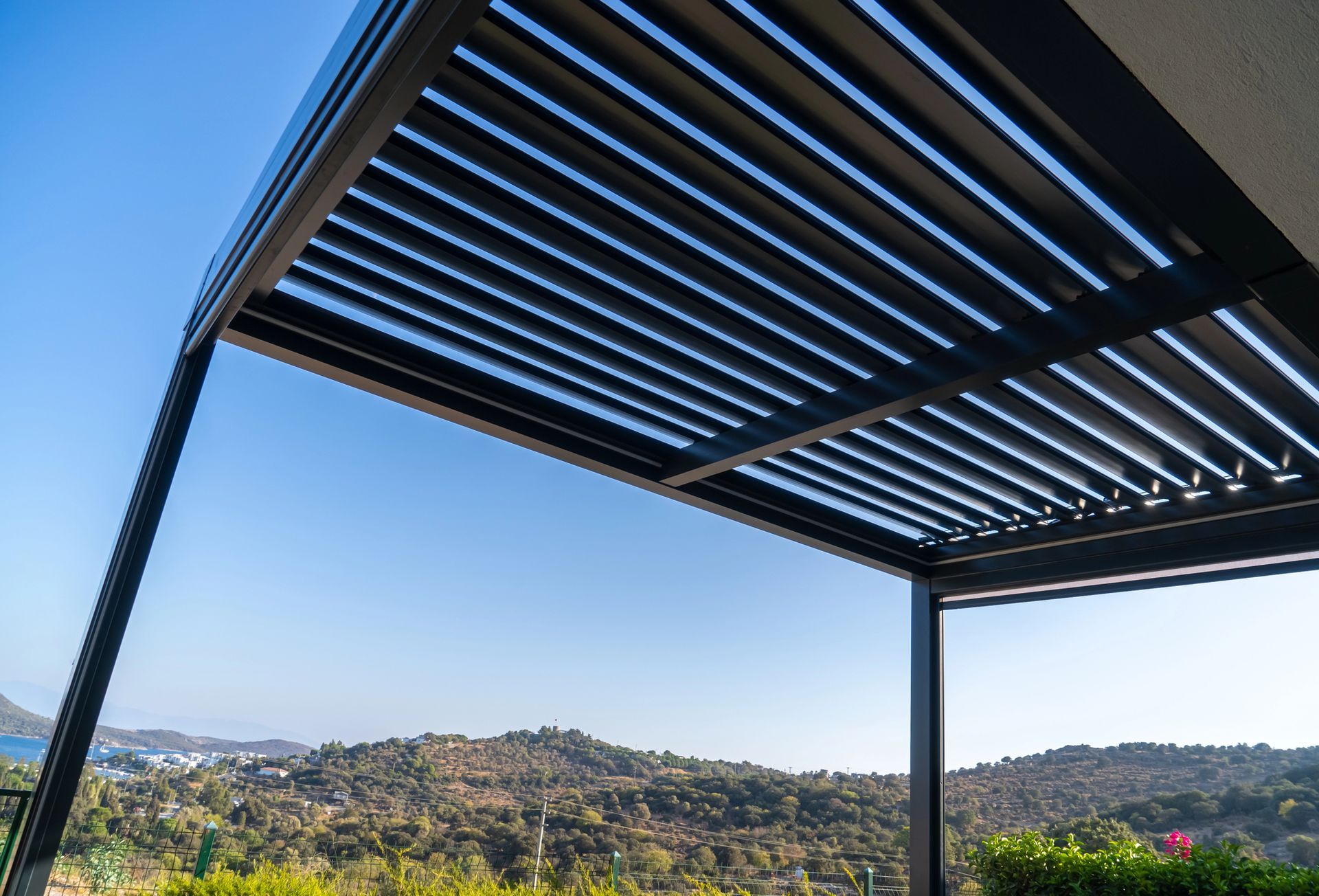 Pergola moderna con tetto a doghe affacciata su una verde collina sotto un cielo azzurro e limpido.