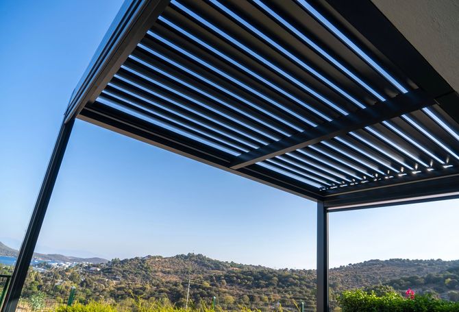 Pergola moderna con tetto a doghe affacciata su una verde collina sotto un cielo azzurro e limpido.
