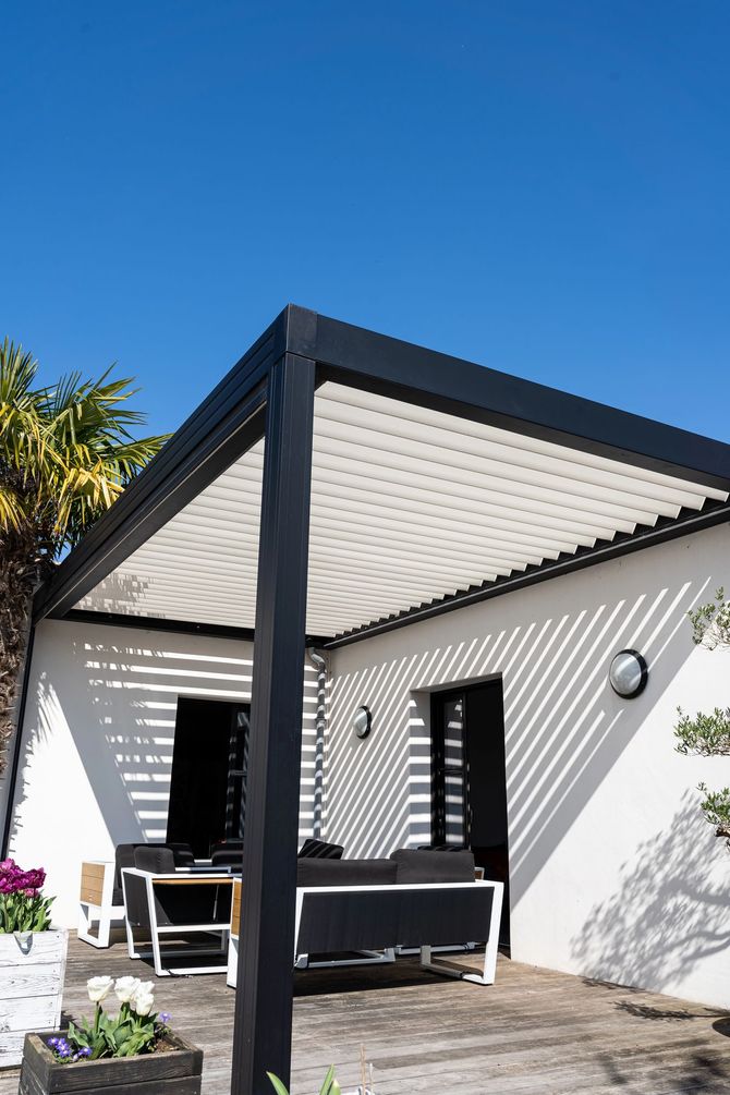 Patio moderno con tettoia dalla struttura nera, pareti bianche e posti a sedere all'aperto sotto un cielo azzurro limpido.