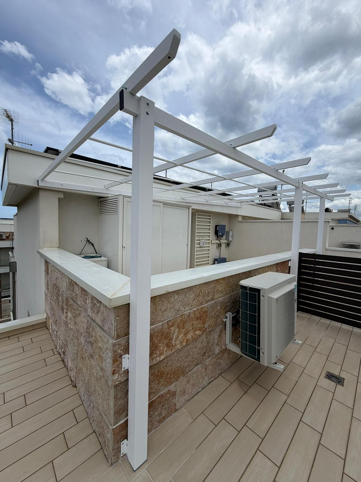 Terrazza sul tetto con pergola bianca, pavimento in legno color sabbia e cielo nuvoloso sopra la casa.