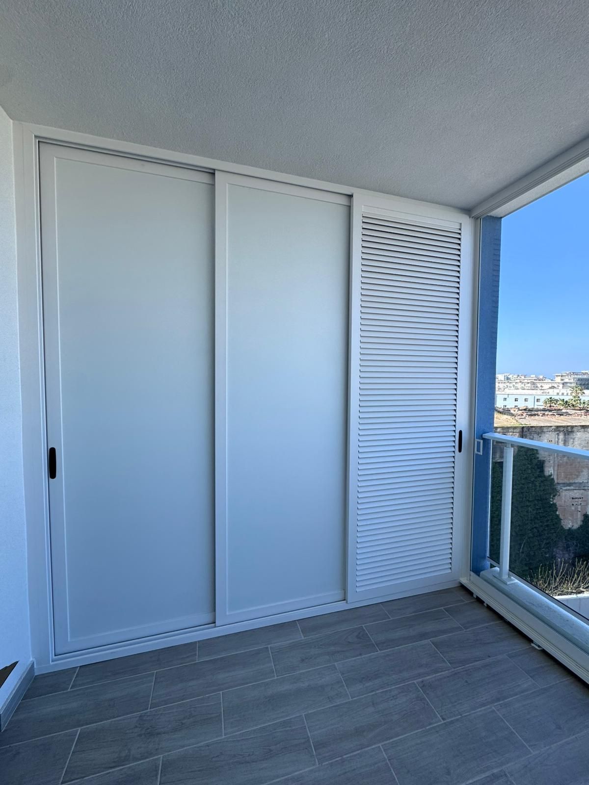 Balcone bianco con ante scorrevoli per l'armadio, pannello a persiana e ringhiera in vetro con vista sulla città.