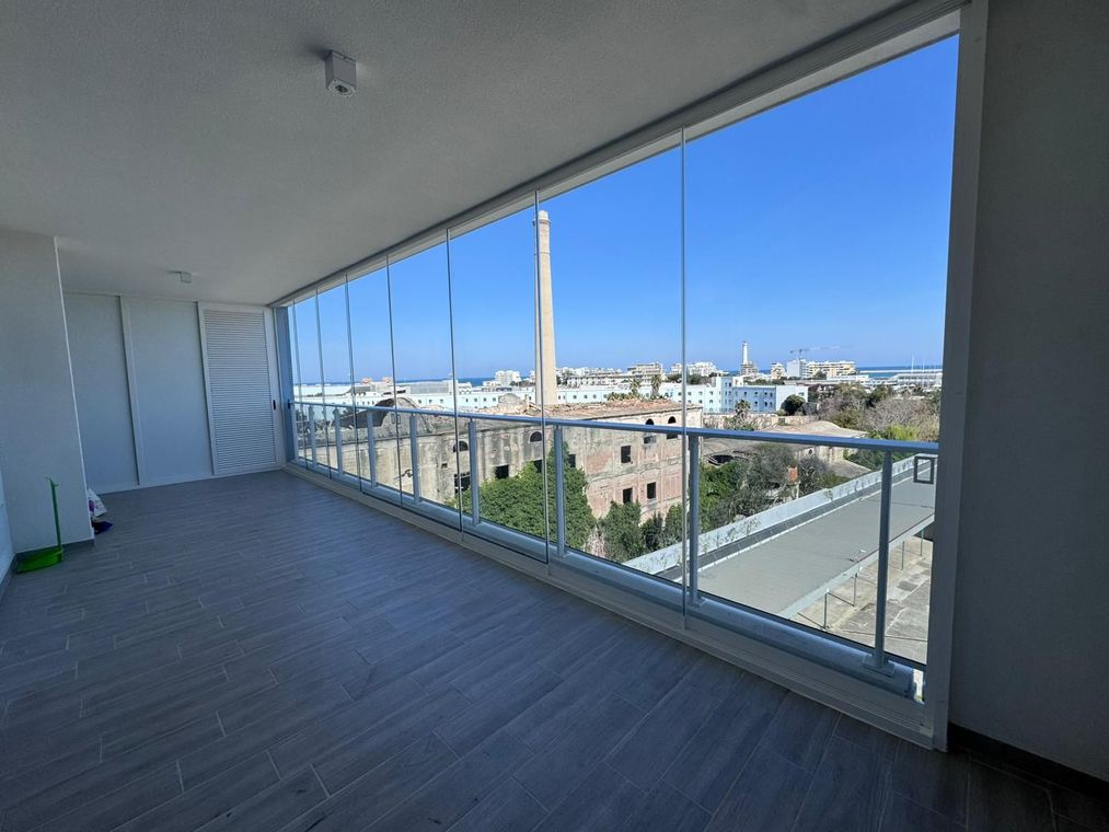 Balcone coperto vuoto con vetrate a tutta altezza affacciate sulla città e sul cielo azzurro.