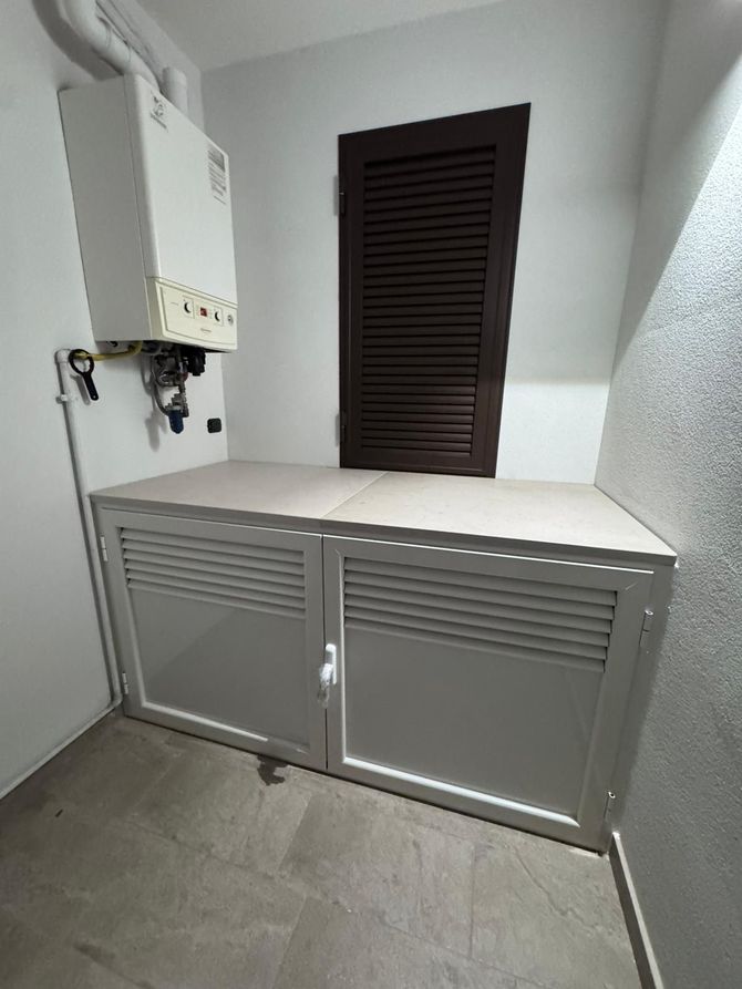 Piccolo locale di servizio con caldaia a muro e armadietti grigi sotto una porta scura a persiana.