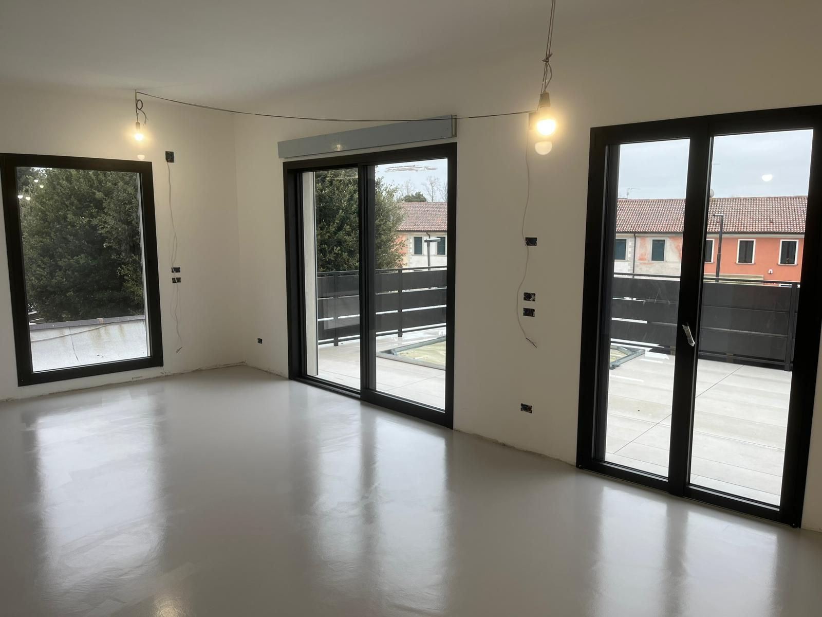 Stanza moderna e vuota con pareti bianche, pavimento lucido e grandi porte finestre con cornice nera che si aprono su un patio.