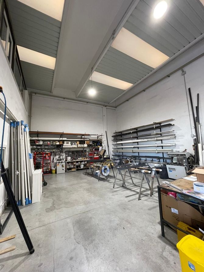 Ampia officina con scaffali in metallo, banchi da lavoro, attrezzi e luminose luci a soffitto.