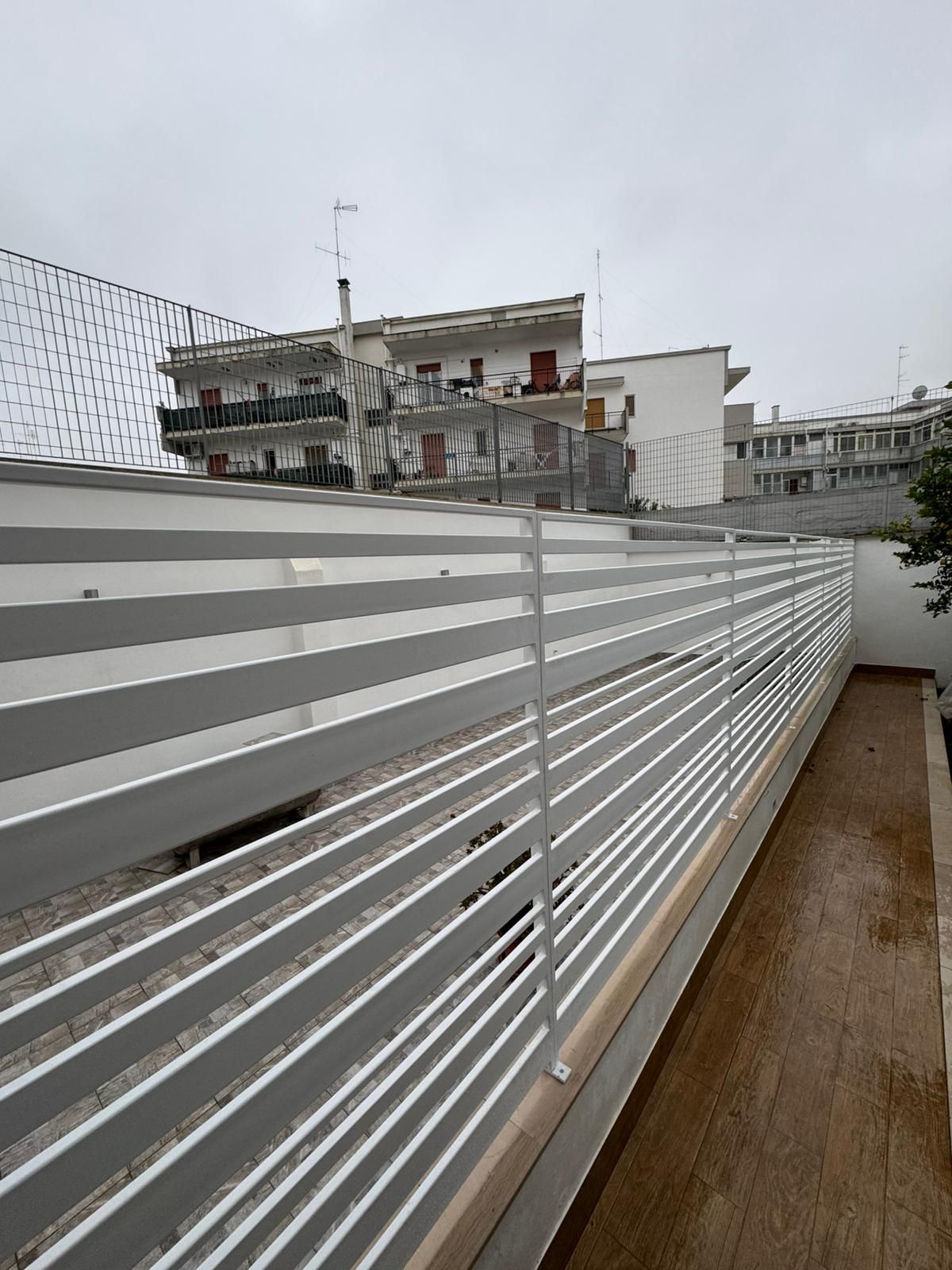 Una moderna recinzione metallica a lato di un passaggio pedonale, con palazzi residenziali sullo sfondo sotto un cielo nuvoloso.