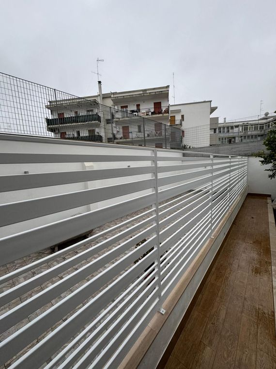 Una recinzione a doghe grigie accanto a un vialetto bagnato, con palazzi sullo sfondo sotto un cielo nuvoloso.