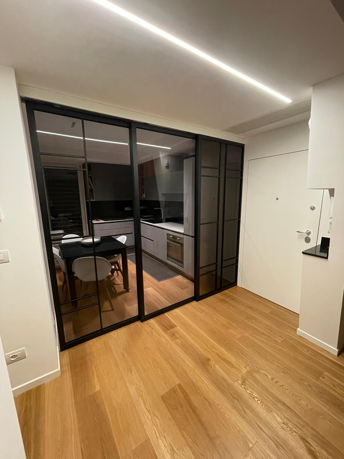 Appartamento moderno con cucina e zona pranzo, pavimenti in legno, ante scorrevoli in vetro nero e illuminazione a LED a soffitto.