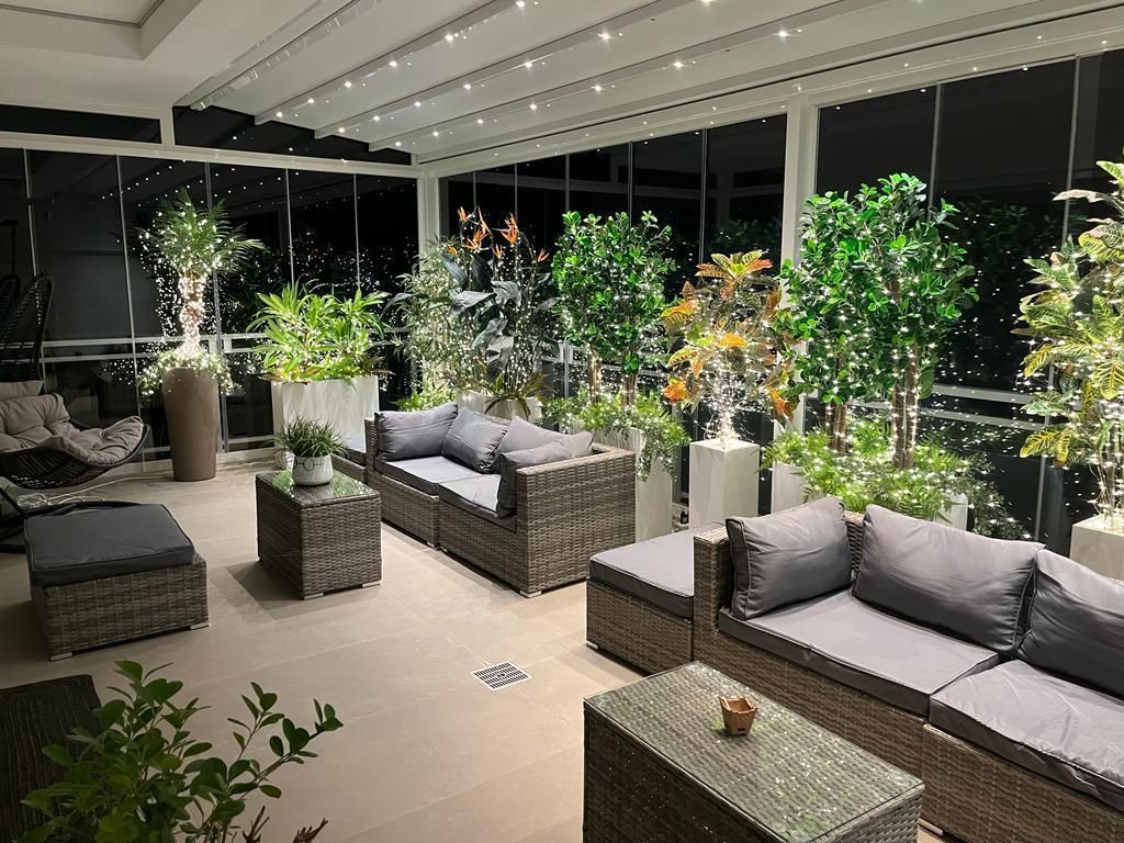 Moderno salotto da esterno con divani in vimini, piante in vaso e luci decorative notturne.