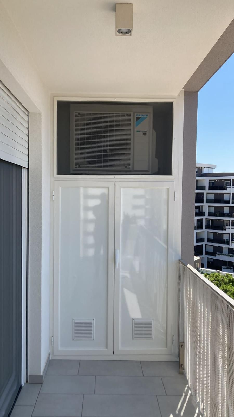Balcone con armadietti bianchi sotto una griglia di ventilazione, accanto a una ringhiera e a palazzi residenziali.