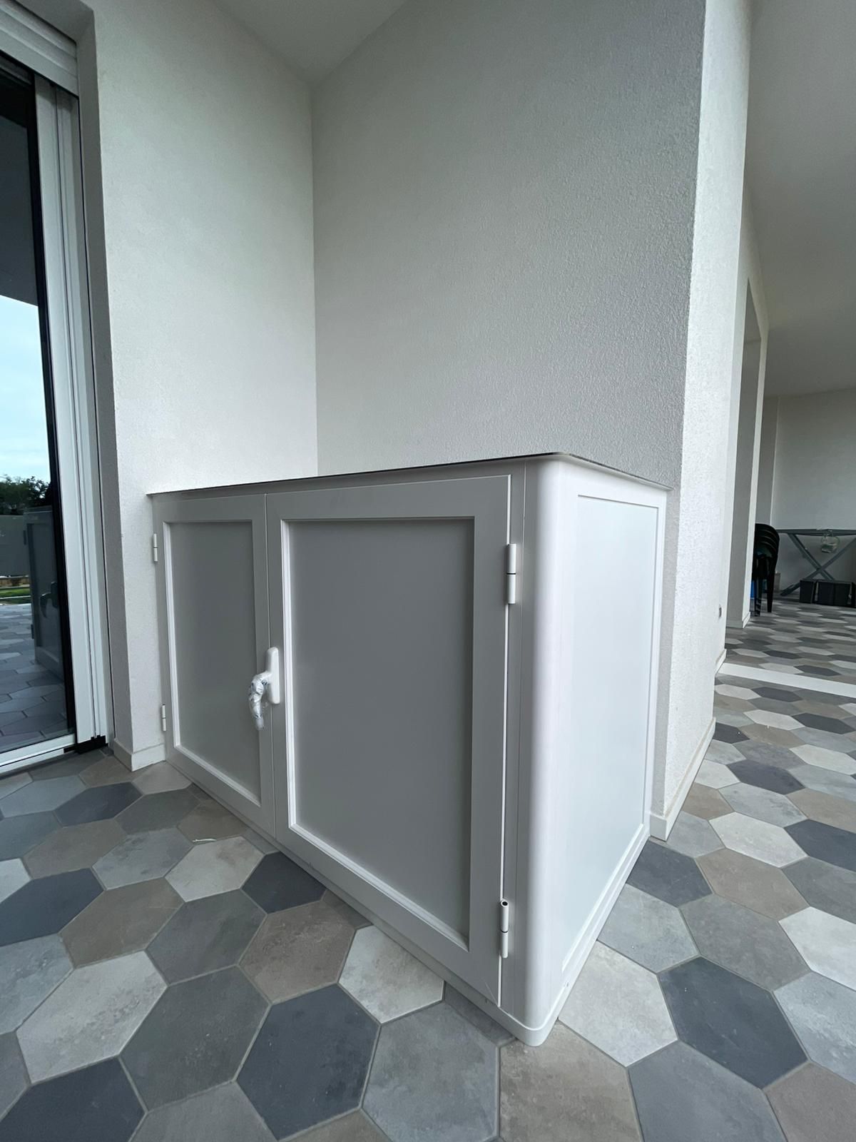 Mobiletto da esterno bianco con due ante su un patio piastrellato vicino a un muro e porta in vetro