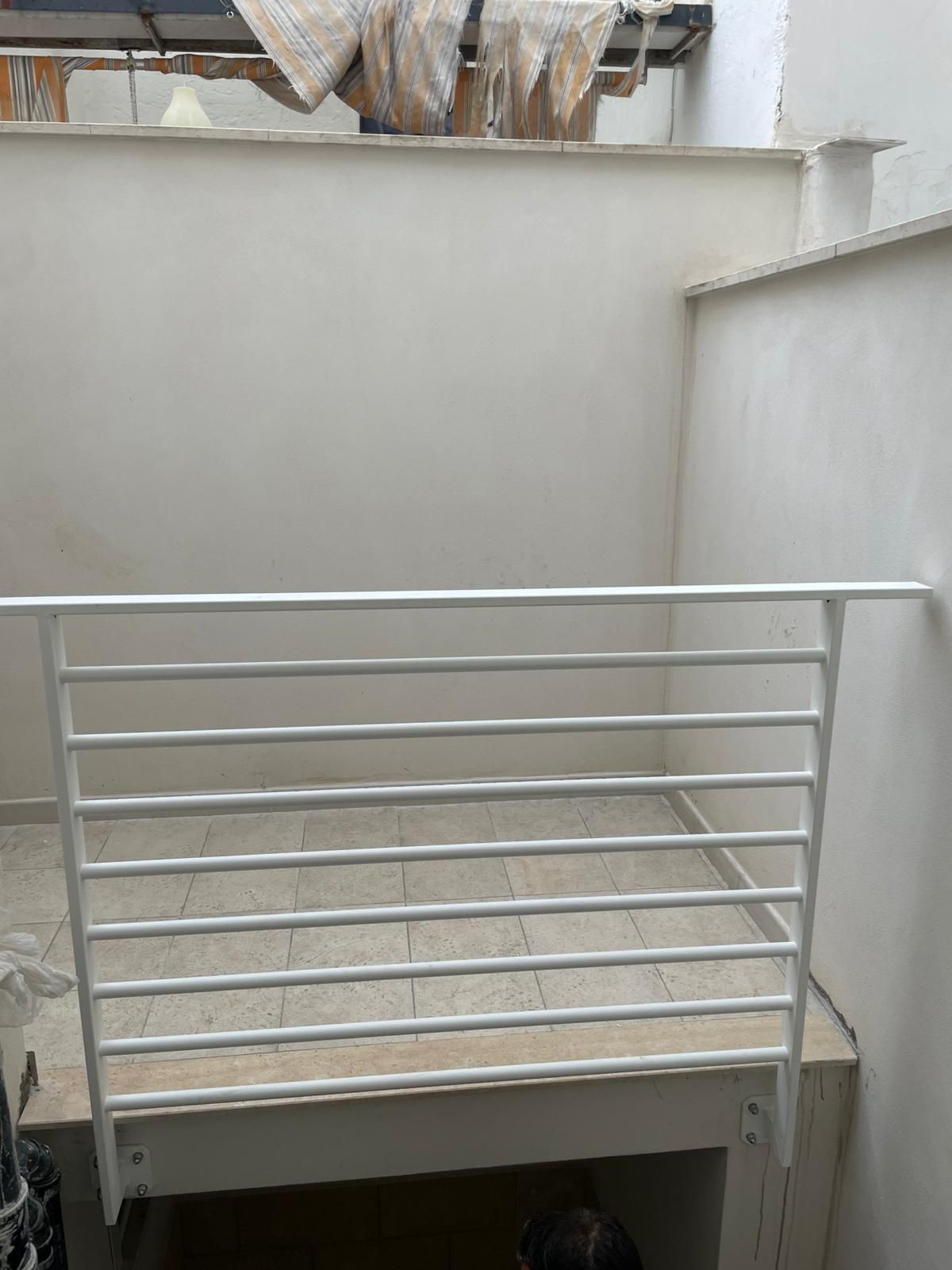 Ringhiera in metallo bianco su un piccolo balcone o pianerottolo, accanto a pareti grigio chiaro e pavimento piastrellato.