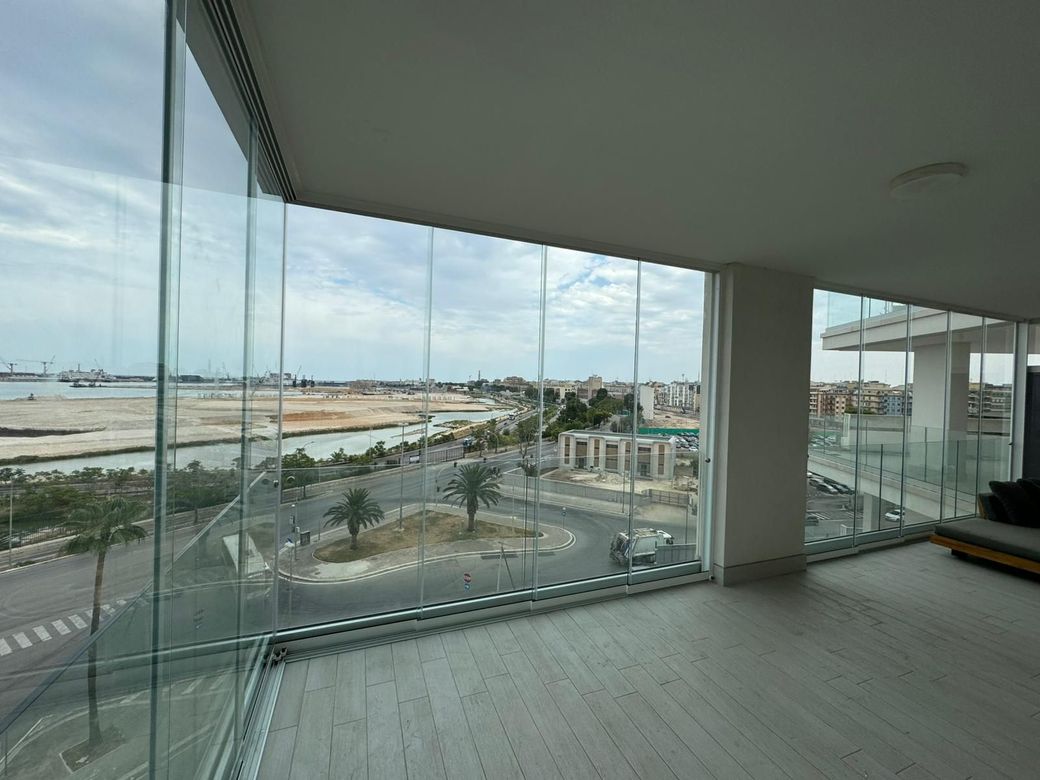 Balcone vuoto con ringhiera in vetro affacciato su una spiaggia e un porto turistico in una giornata nuvolosa.