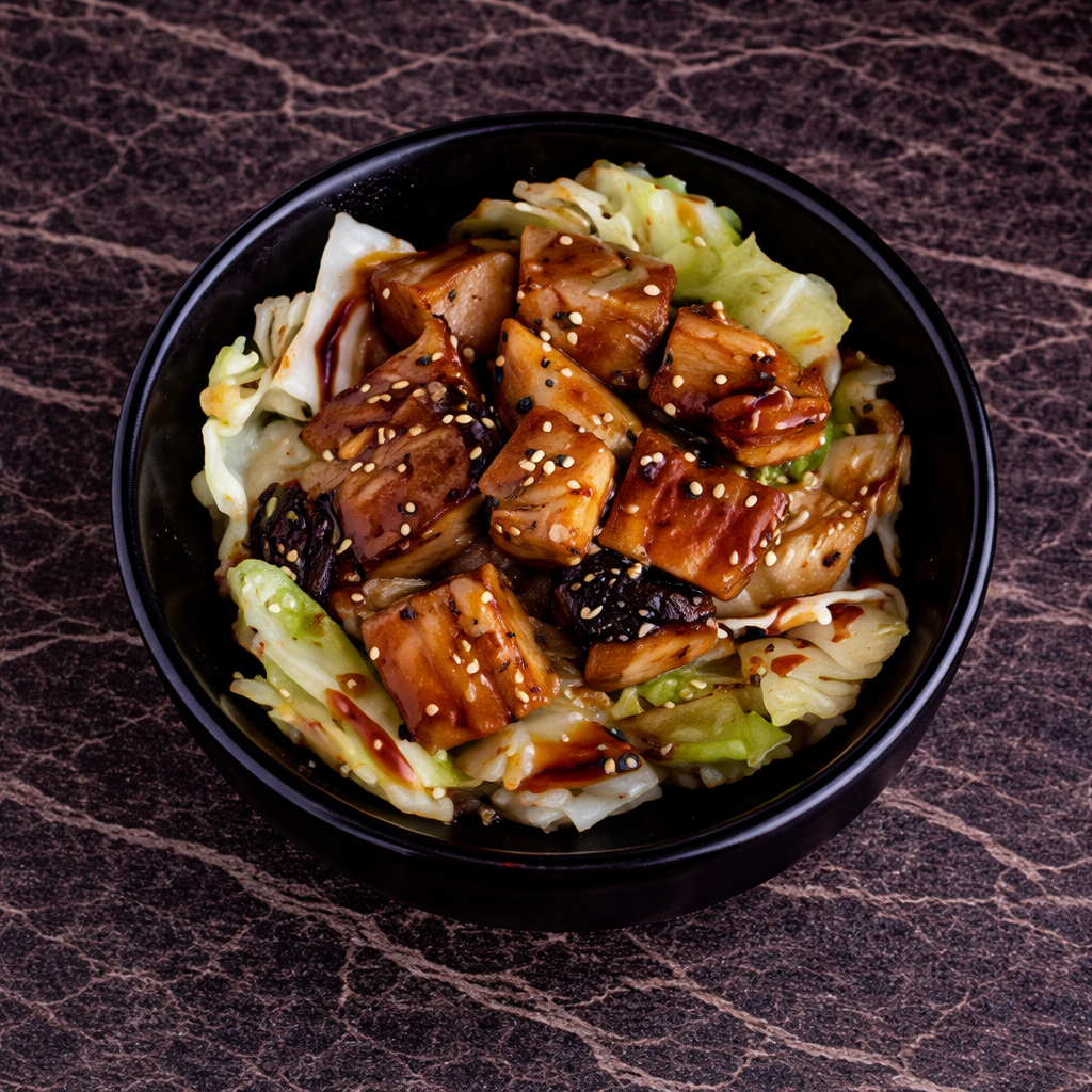 A bowl of glazed, cubed protein pieces served over a bed of sautéed cabbage, sprinkled with sesame seeds on a dark table.