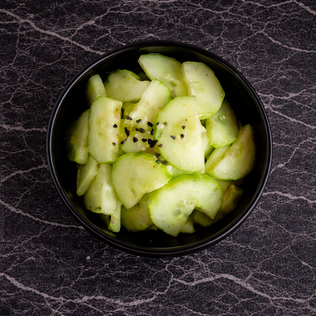 A small black bowl filled with sliced cucumber salad garnished with black sesame seeds, set on a dark textured surface.
