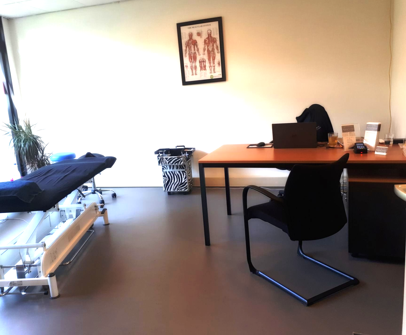 Fysiotherapiepraktijk met onderzoekstafel, bureau en een anatomische kaart van het menselijk lichaam aan de muur.