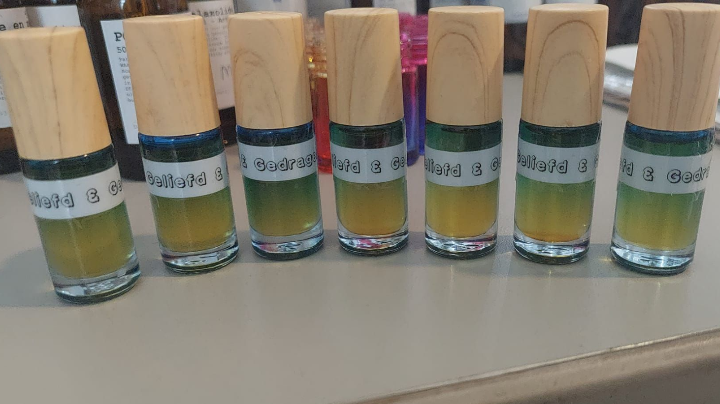 Zes glazen flessen met houten doppen, gevuld met gelaagde gele en groene vloeistoffen, voorzien van tekstetiketten.