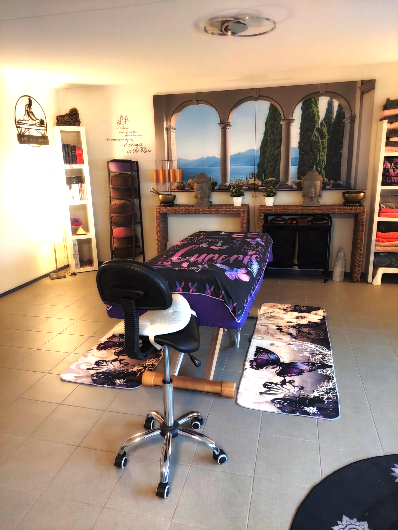 Massagetafel met zwart laken met bloemenpatroon, rolkussen en een zachtgrijs vloerkleed.