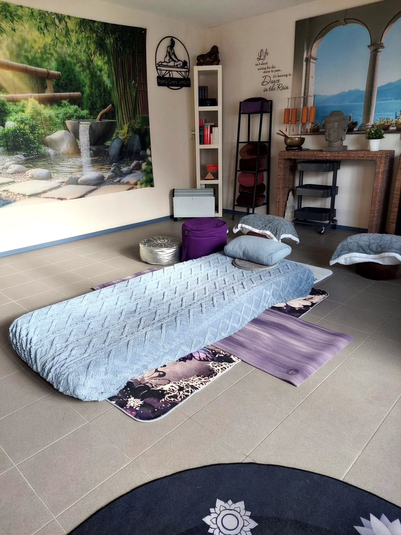 Ontspanningsruimte met massagetafel, paarse poef en decoratieve plank.
