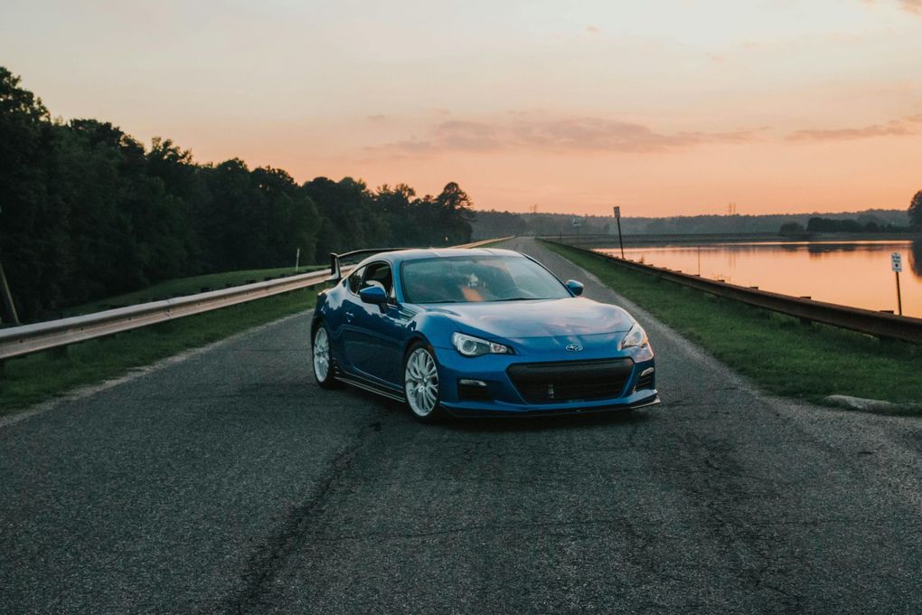 A blue car is driving down a road next to a body of water.