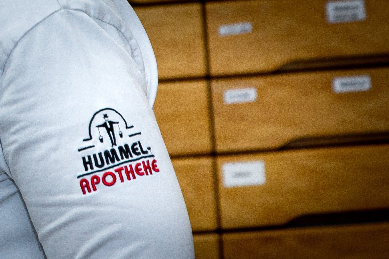 Vor einer Schubladenreihe steht eine Person, die ein Hummel-Apothekenhemd trägt.