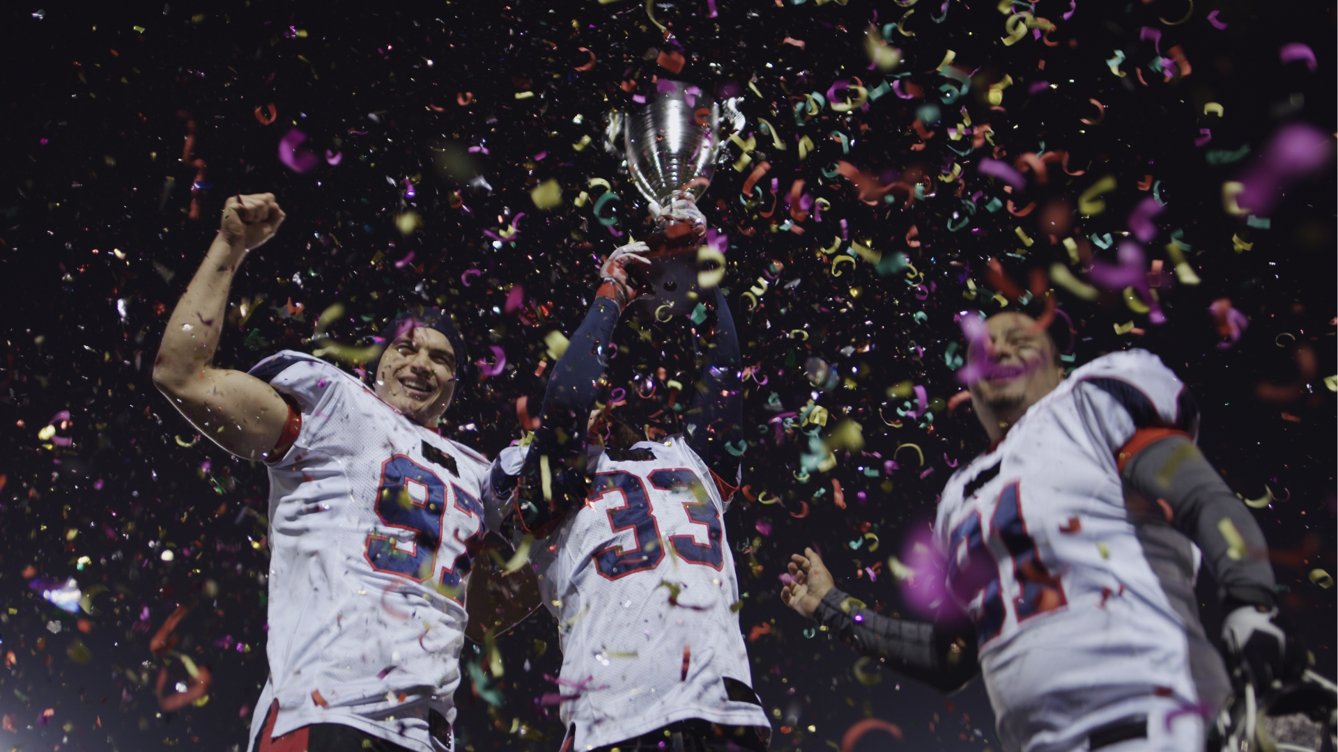 Three football players are holding a trophy and confetti is falling around them.
