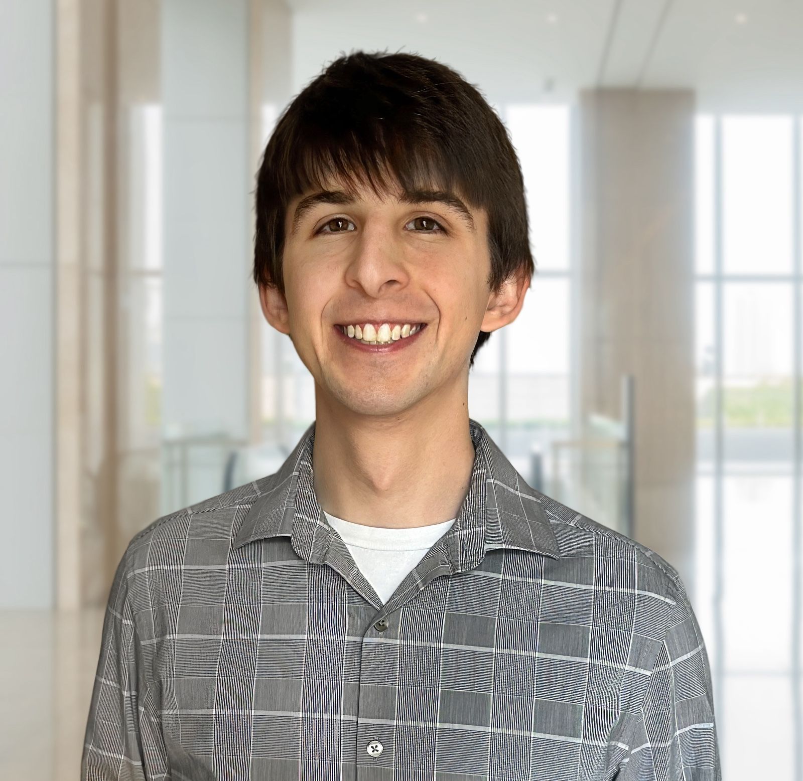 Man with dark hair and a patterned shirt smiles in a bright indoor hallway.