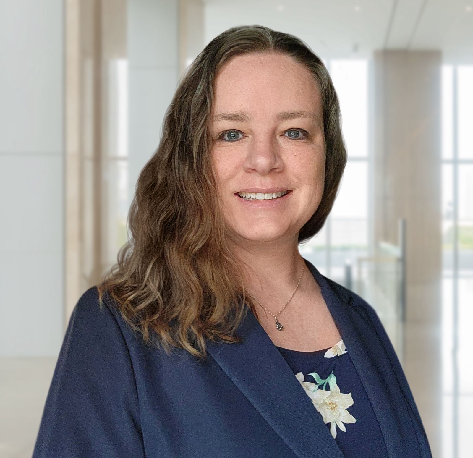Woman with shoulder-length brown hair, in a navy blazer, smiles in a modern, light-filled office.