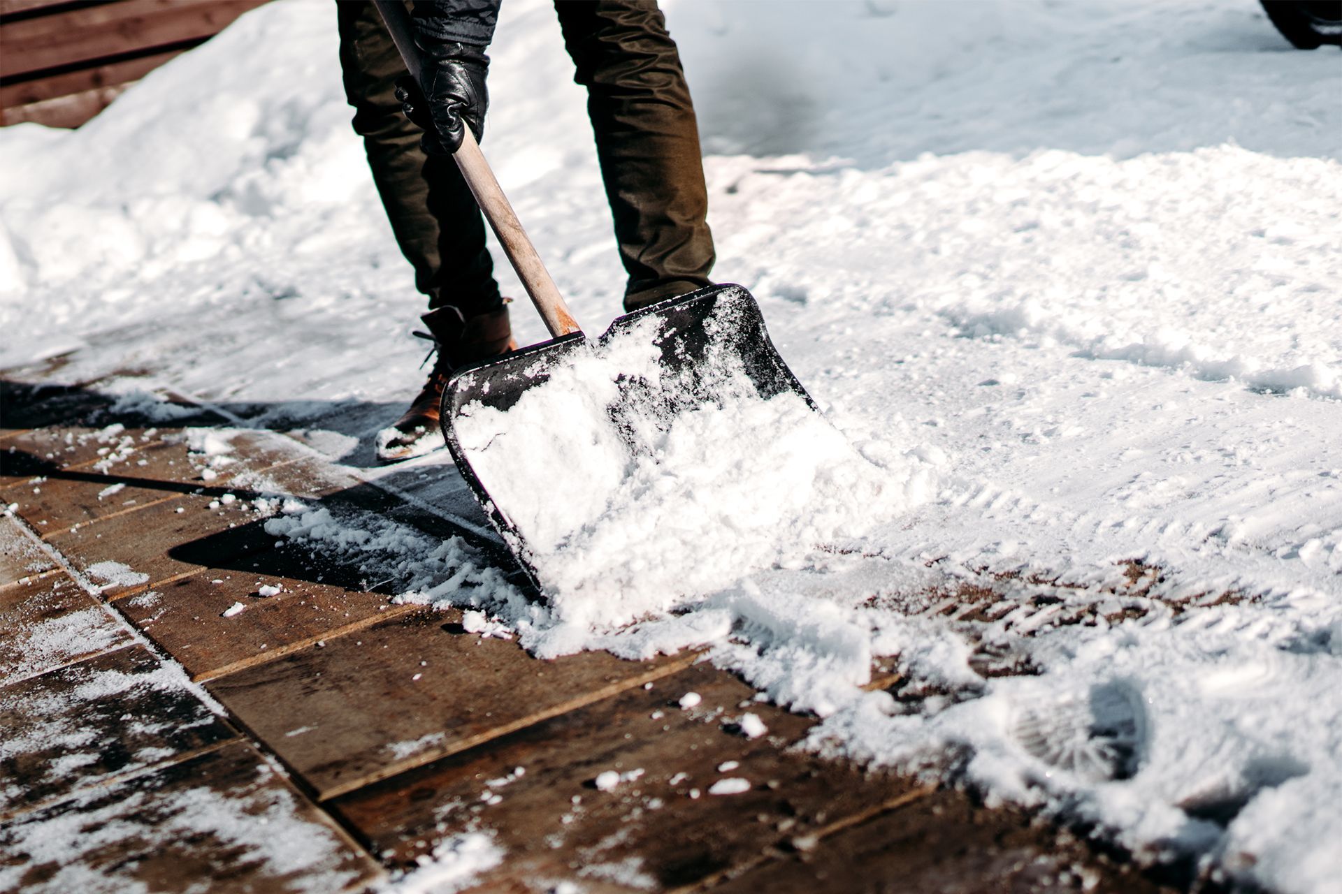 Person shoveling snow from a wooden deck with a black shovel on a sunny day.