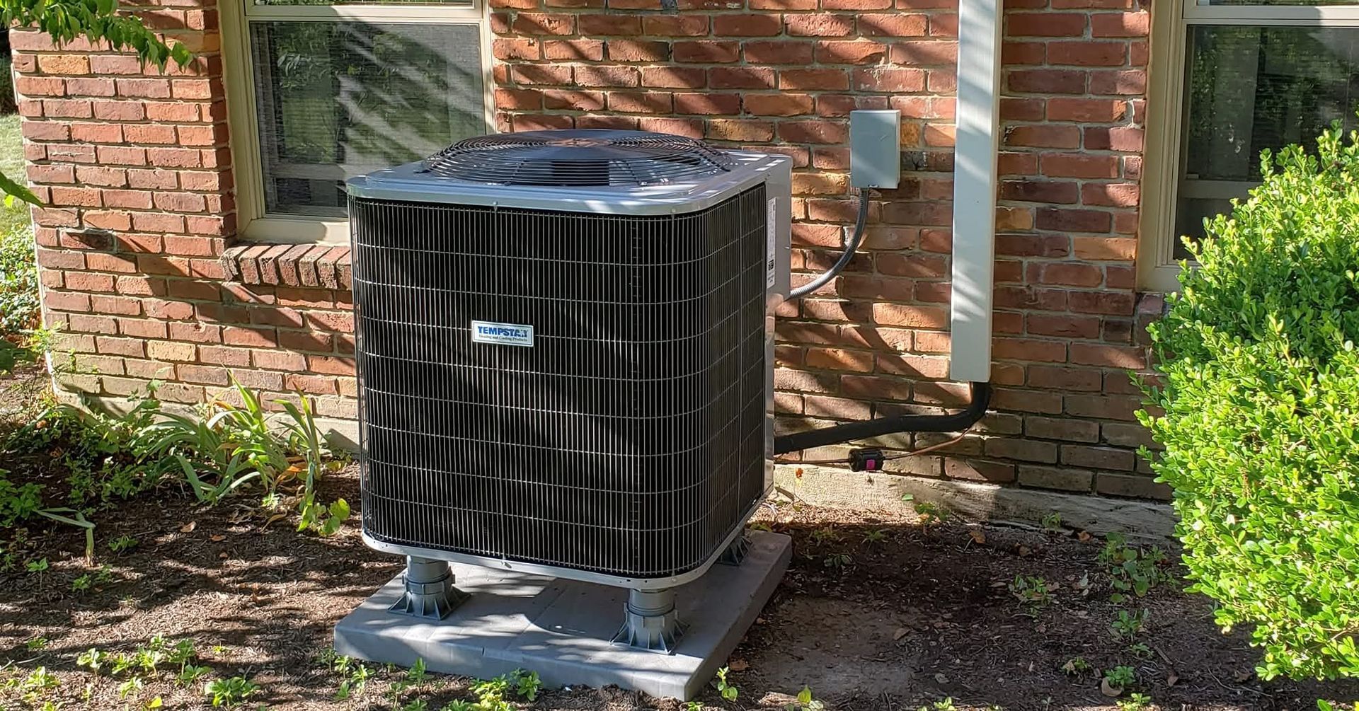 Central air conditioner unit against a brick wall, next to a bush.