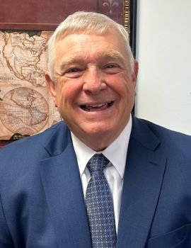 Smiling man in a blue suit and tie, indoors with a map on the wall behind him.