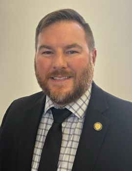 A man in a suit with a tie and a beard smiles at the camera.