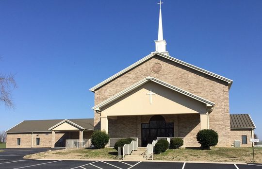 Church building with beige brick exterior, white trim, and a cross on top.