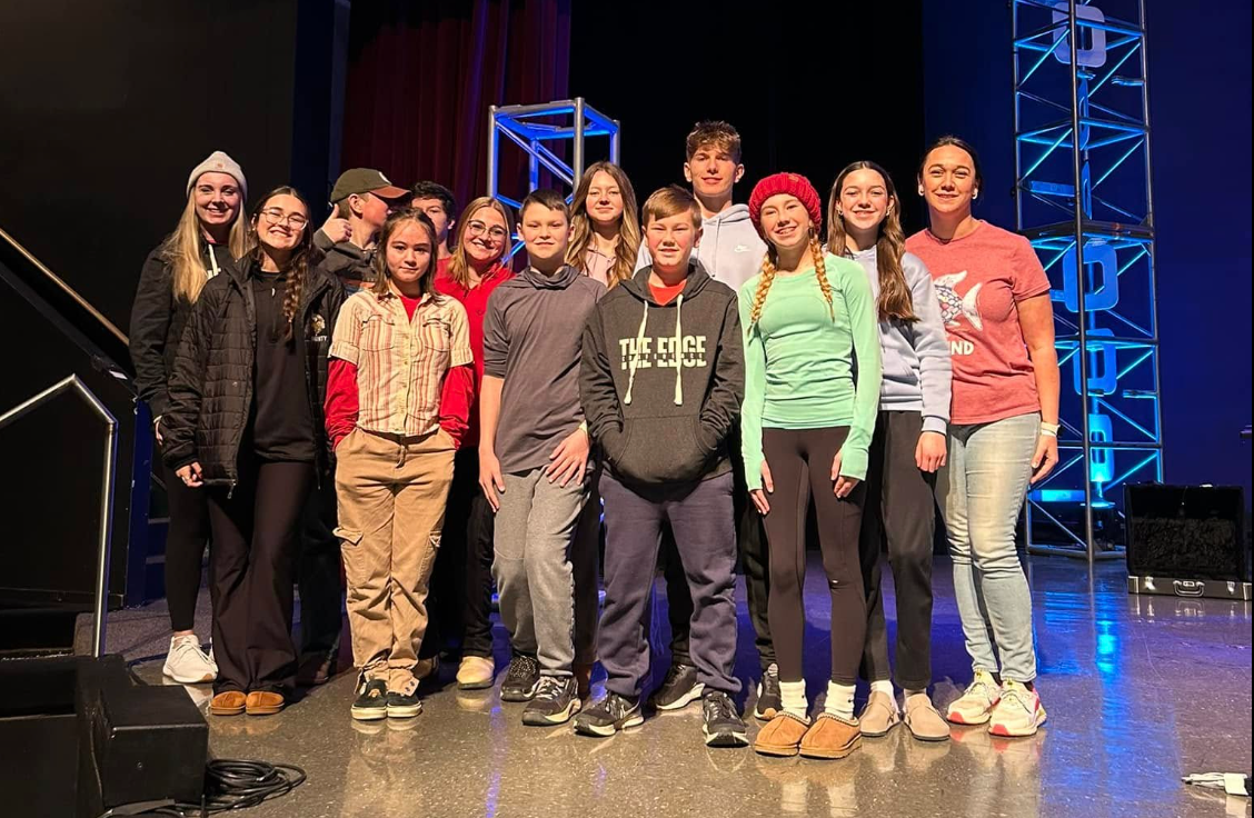 A group of teens on a stage, smiling and posing, with a red curtain backdrop.