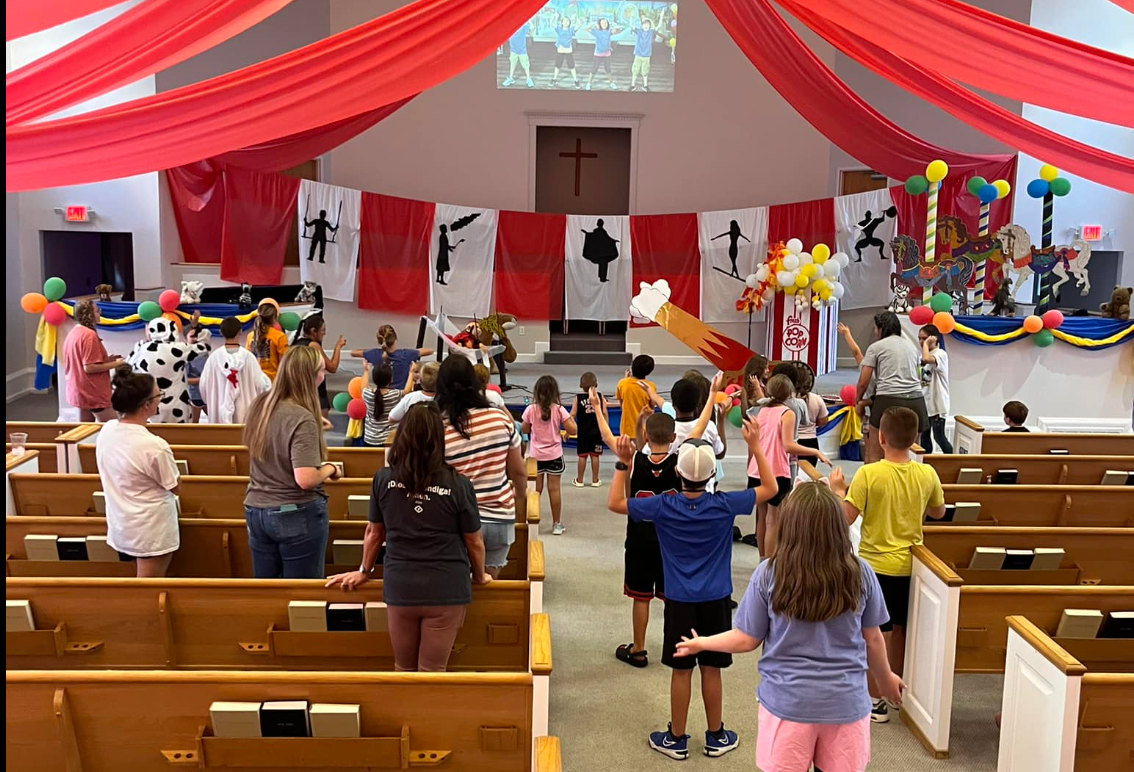 Children in a church event with red decorations and balloons. People standing and raising arms.