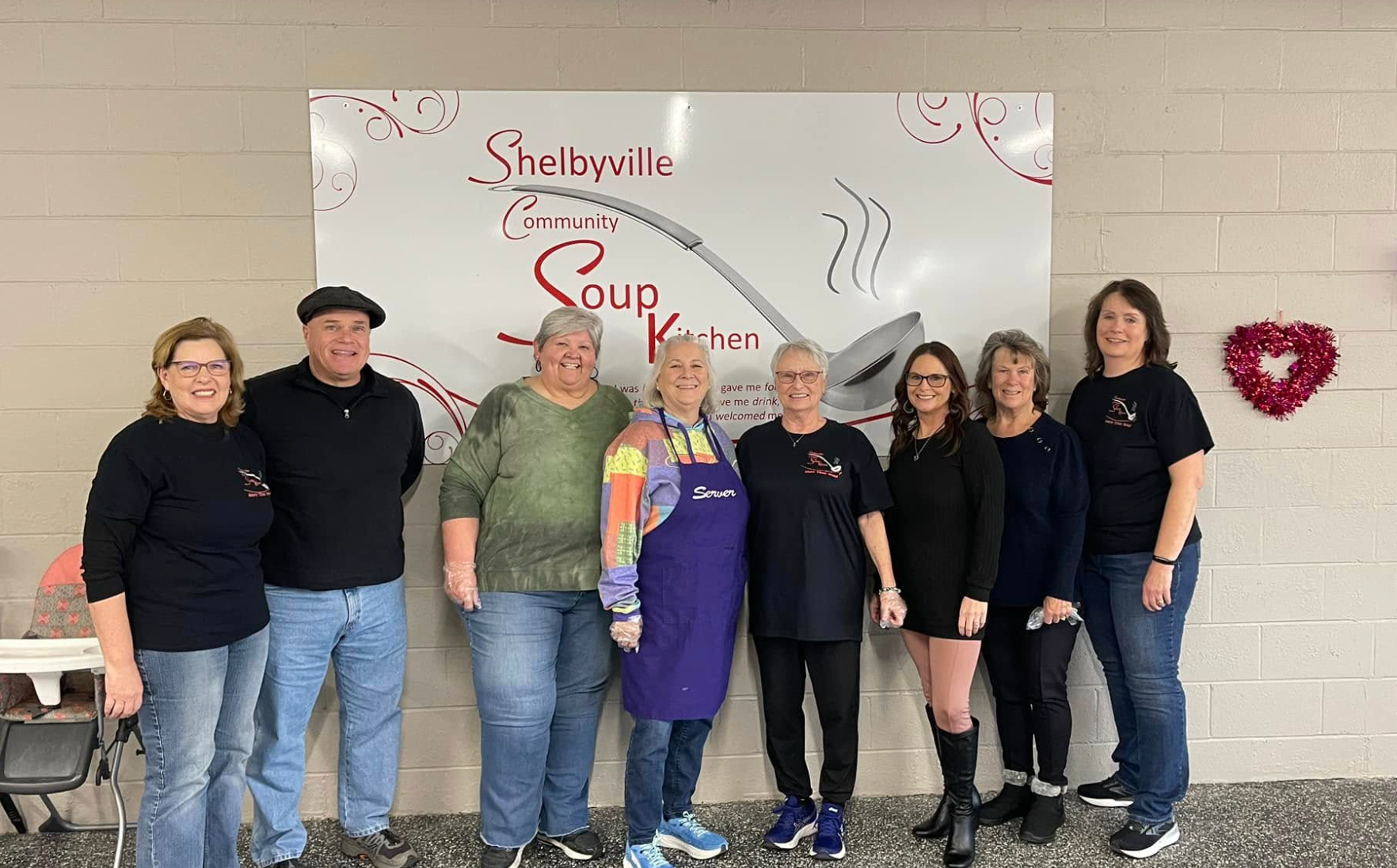 Shelbyville's Corner Soup Kitchen staff posing with sign. Smiling people of various ages, inside.