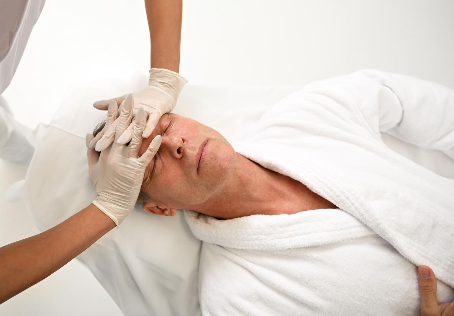 A professional in gloves performs a facial massage on a person lying down in a white spa robe.
