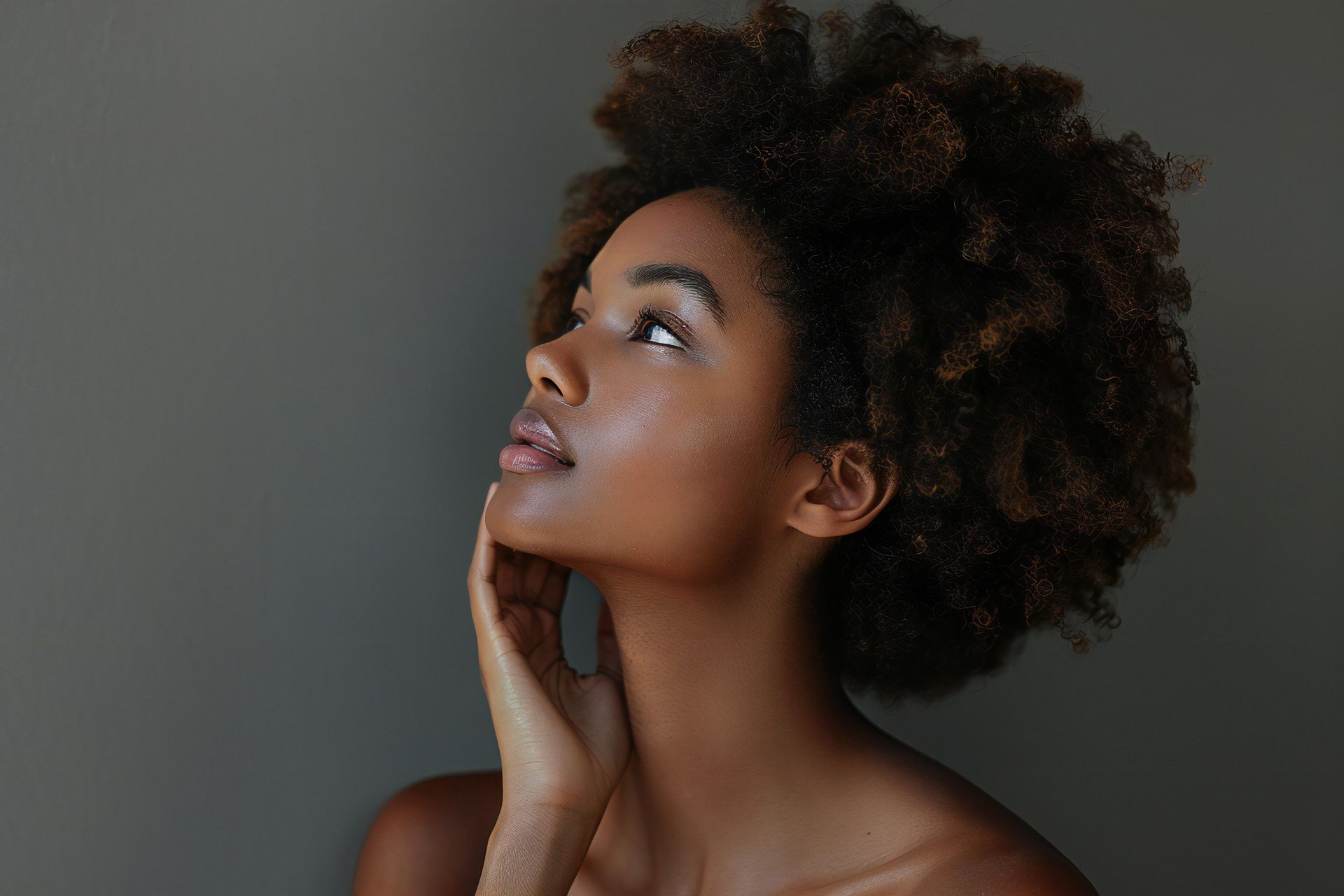 Person with dark skin looks up, hand on neck. They have natural, curly hair against a gray background.