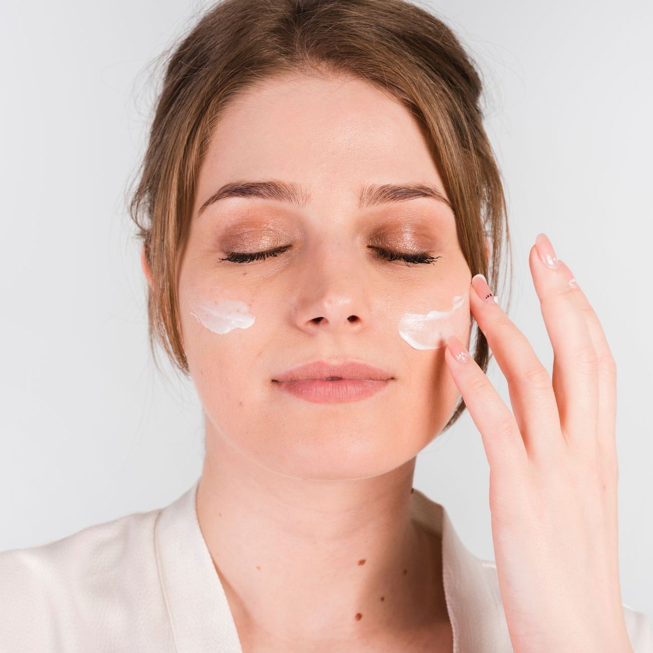A woman is applying cream to her face with her eyes closed.