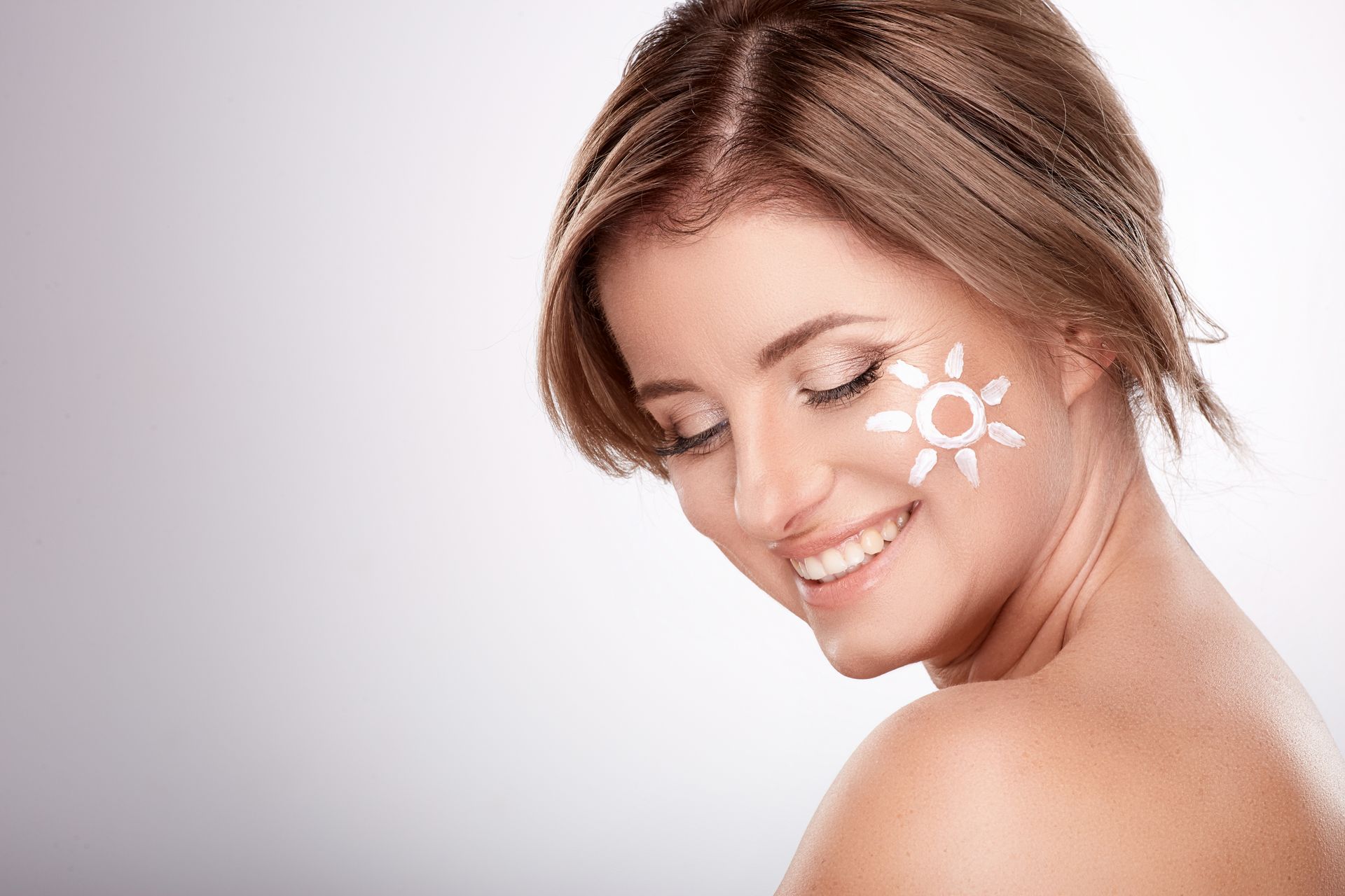 Woman with sunscreen sun on her cheek smiling; light background.
