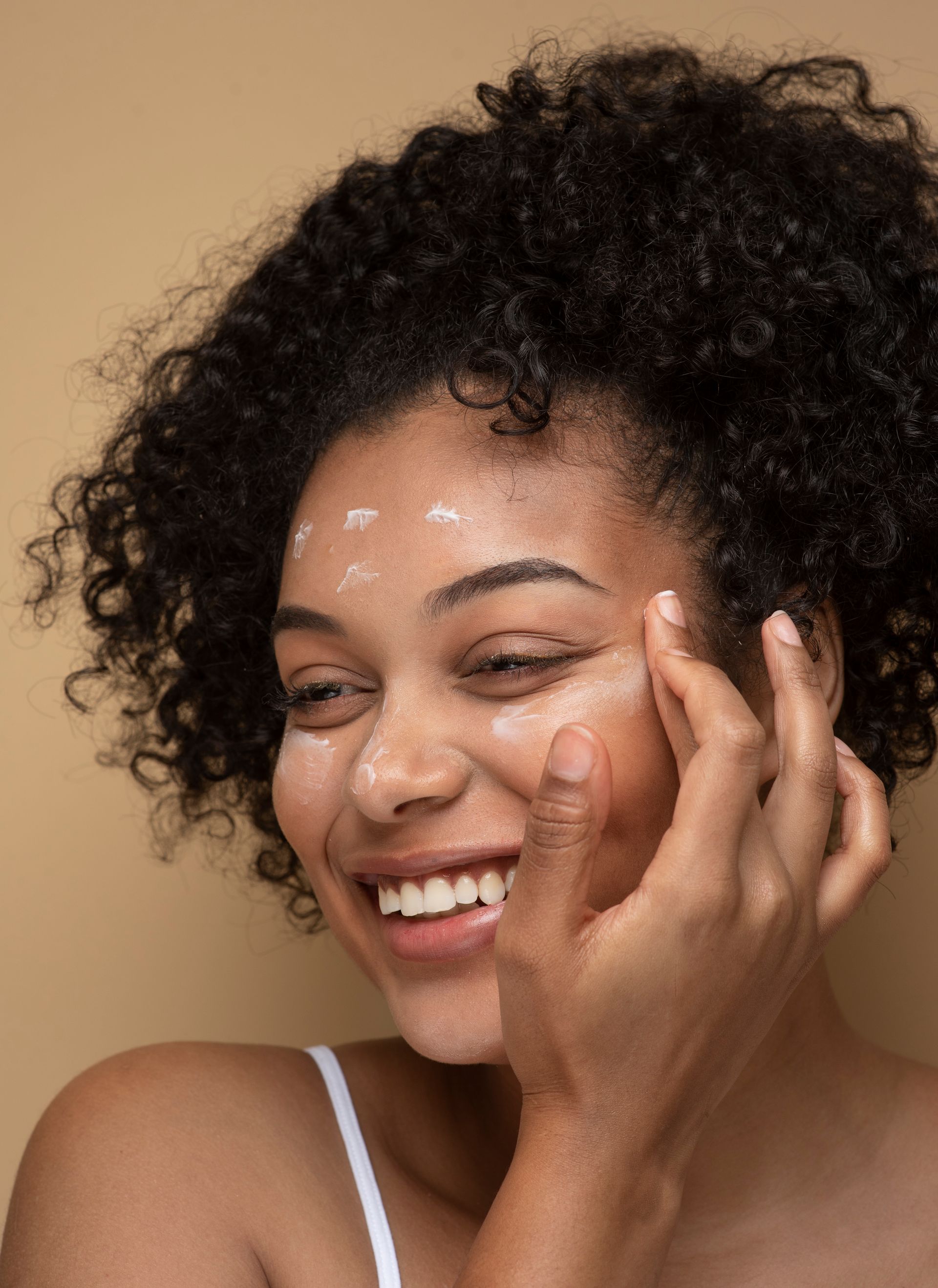 A woman with curly hair is smiling while applying cream to her face.