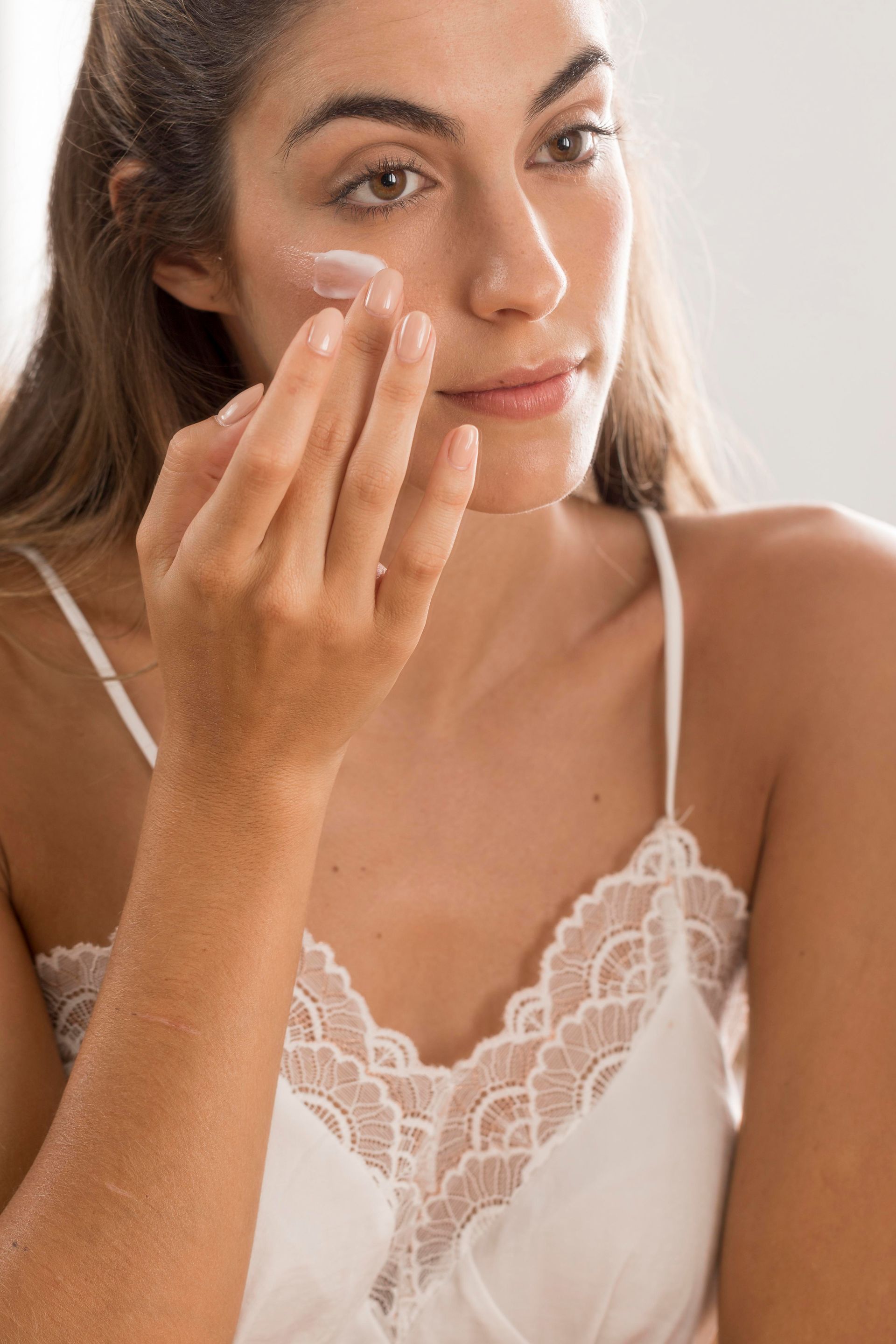 A woman in a white tank top is applying cream to her face.