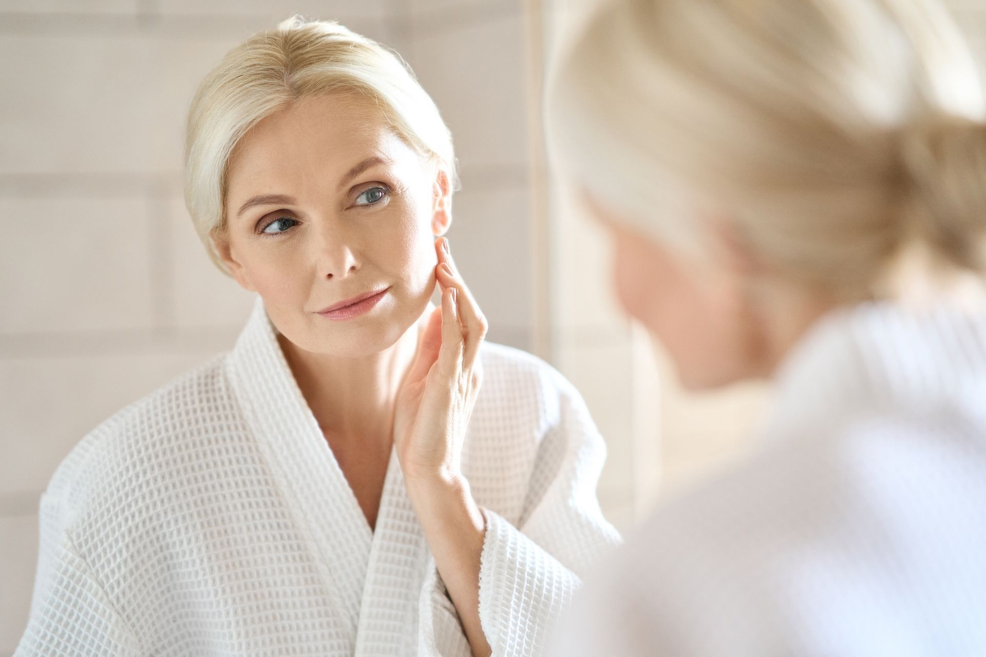 Woman in white bathrobe looking in a mirror, touching her face. Brightly lit bathroom.