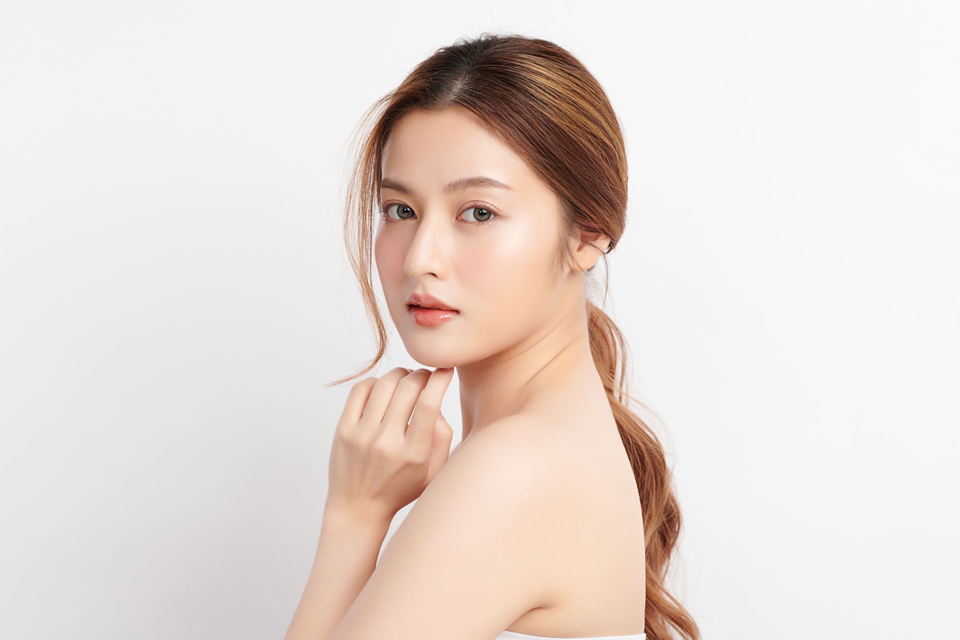 Woman with fair skin and brown hair, looking over her shoulder, hands near face, white background.
