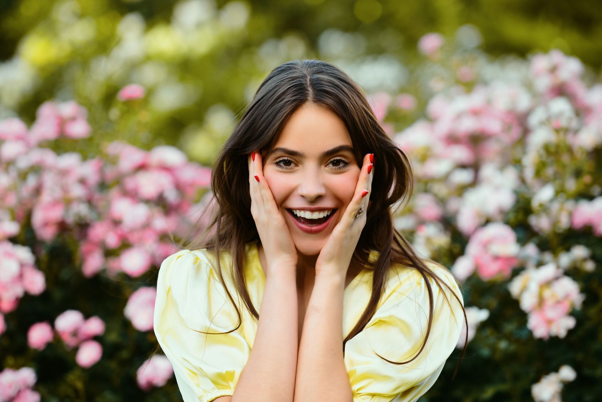 Woman in yellow top smiles with hands on cheeks, surrounded by pink flowers.
